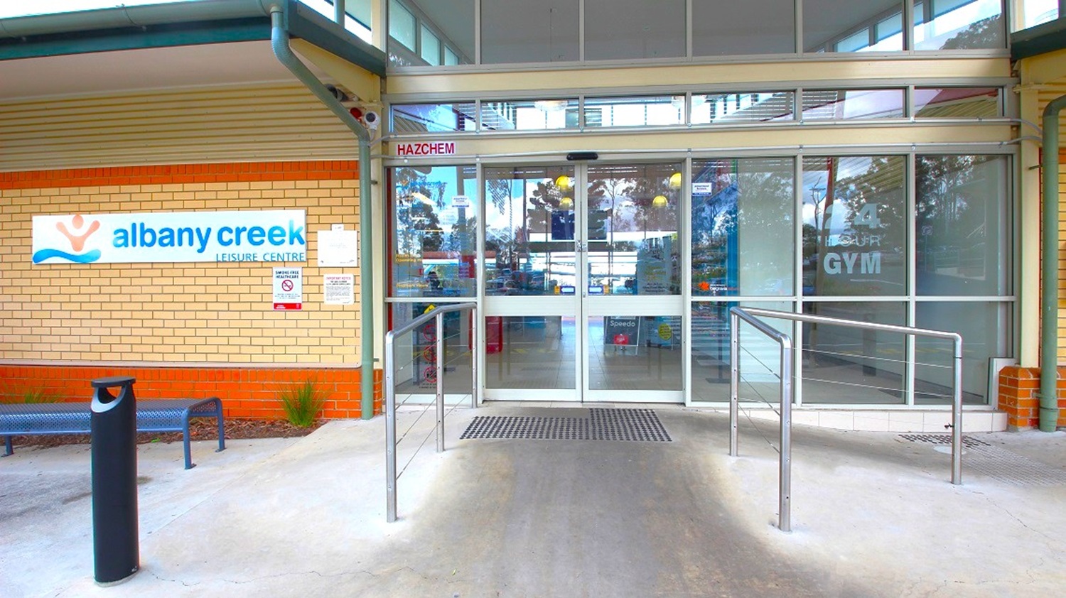 Entrance to a leisure centre with a glass sliding door in the centre. To the left, a brick wall displays a sign reading "albany creek leisure centre" with a bench and a small plant nearby. The right side has a glass panel with "GYM" visible. A metal railing leads up to the entrance, and a mat is placed at the door. The ceiling is glass-panelled, allowing natural light inside.