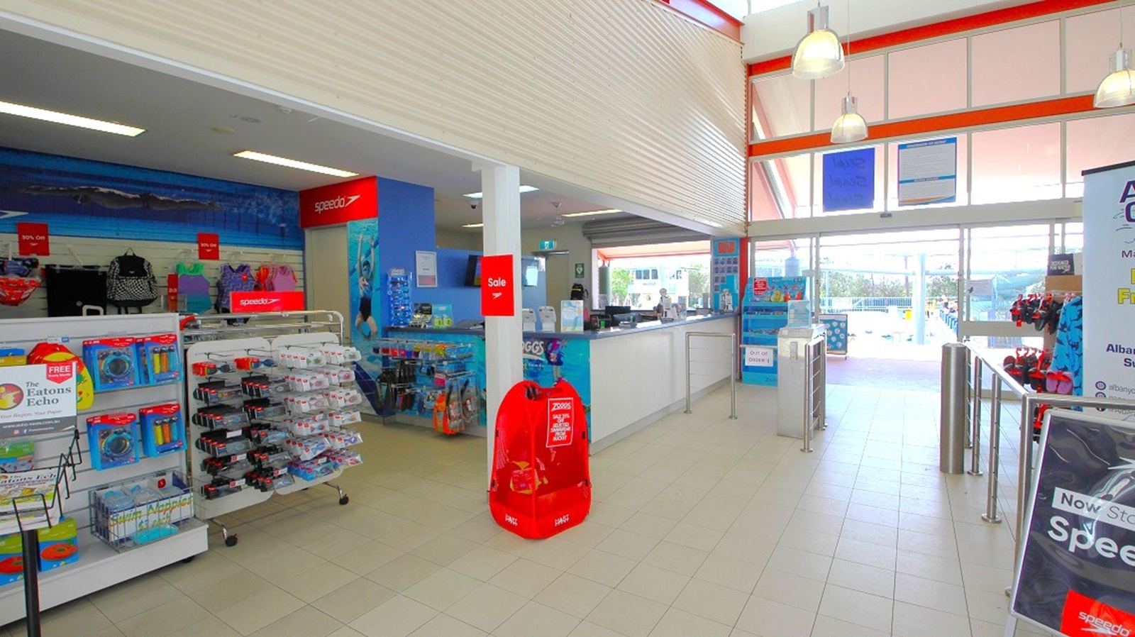 Interior of a shop with a tiled floor and high ceiling. On the left, shelves display various products like swimming gear and accessories. In the centre, a counter with a cash register is visible. To the right, a turnstile leads to a pool area outside, visible through large windows. Bright lighting from ceiling fixtures and natural light from the windows illuminate the space. A red sale sign is attached to a pillar near the centre.