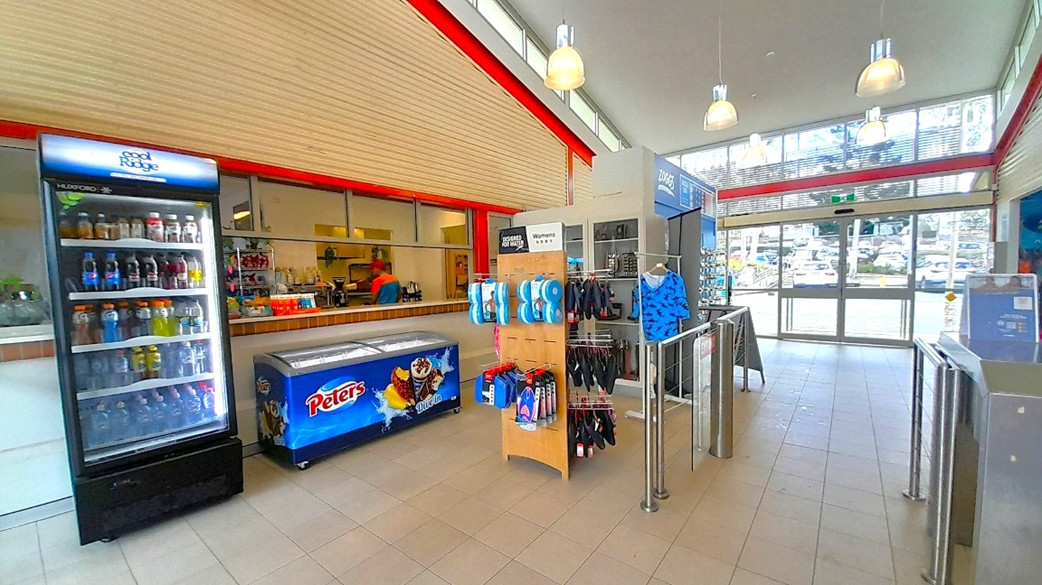 A shop interior with a tiled floor and a high ceiling featuring hanging lights. On the left, there's a refrigerated display filled with drinks and a freezer with "Peters" branding. In the centre, a stand displays various items like swimwear and accessories. The right side has a glass entrance with a view of parked cars outside. The back wall has a counter with snacks and drinks, and a person is visible behind it.