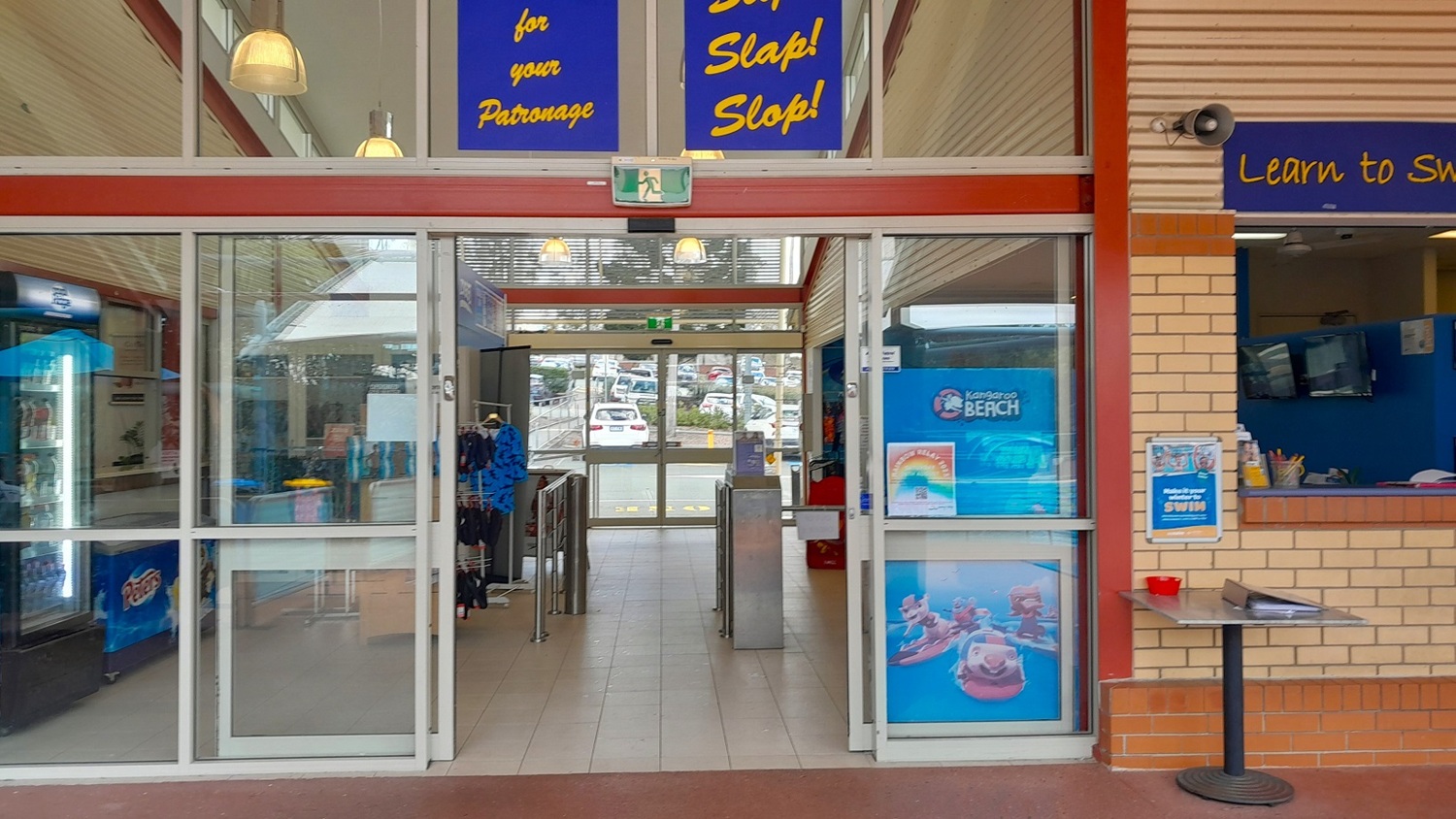 A glass entrance with sliding doors leads into a building. Above the doors, signs read "Slip! Slap! Slop!" and "for your Patronage." Inside, a corridor with tiled flooring extends towards a car park visible through another set of glass doors. On the right, a brick wall features a service window with a blue interior and a small table with a red container. On the left, a display with hanging items and a vending machine is visible. Ceiling lights hang above.