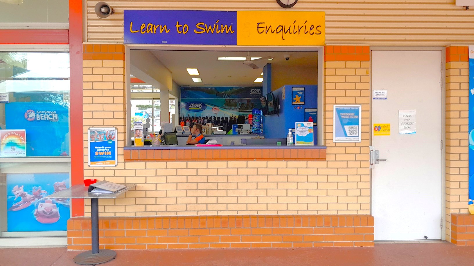 A brick wall with a service window in the centre, labelled "Learn to Swim Enquiries" in yellow and blue. Inside, a person is seated at a desk with a computer. To the left, a window displays a poster with "Kangaroo Beach" and colourful images. Below, a small round table holds a red item and papers. On the right, a white door with notices is visible. The floor is tiled in a reddish-brown colour.