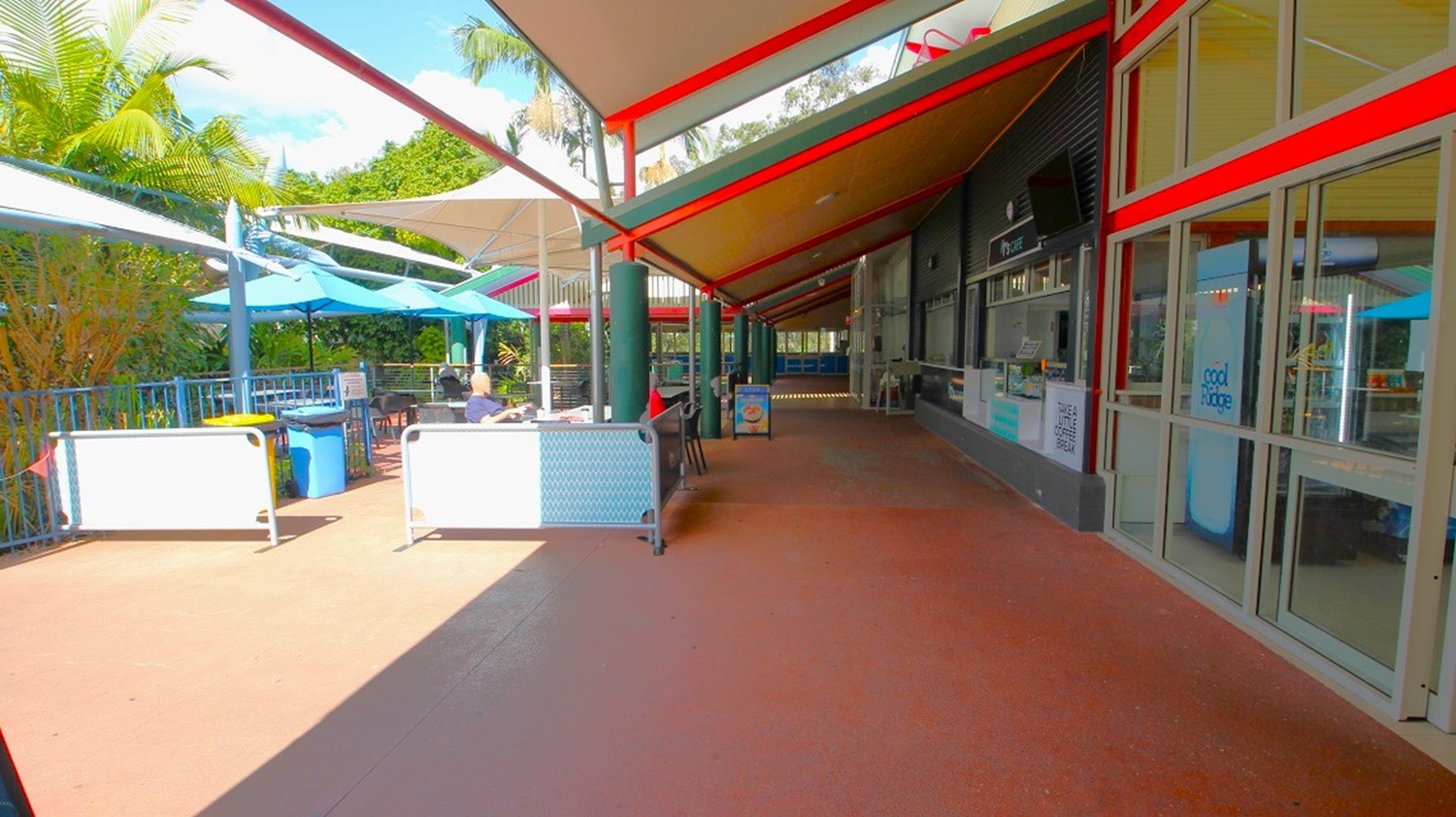 A covered outdoor walkway with a red and beige canopy overhead. On the right, there are glass doors and a wall with signs and a menu. On the left, there are tables with blue umbrellas and people seated, surrounded by greenery. The floor is a reddish-brown tile, and there are metal railings along the left side.