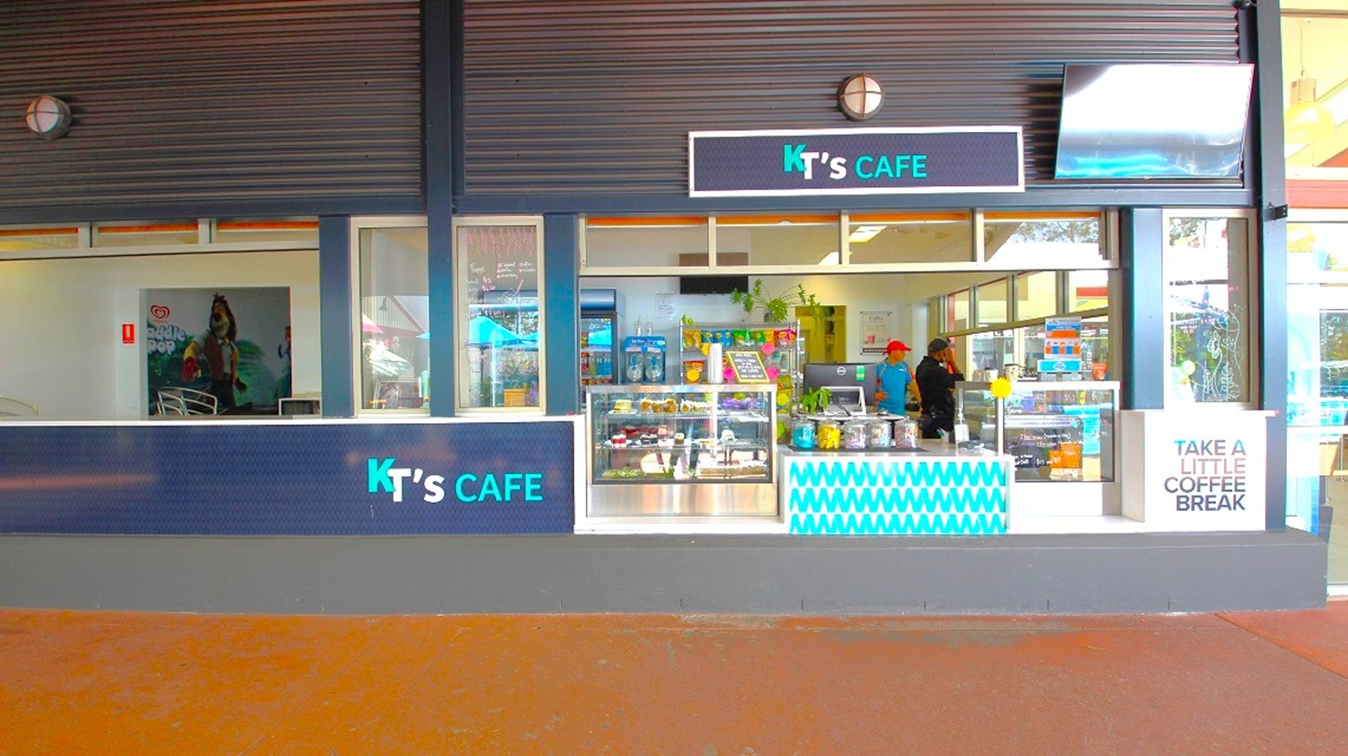 A café storefront with a sign reading "KT's Café" above the entrance. The front features large glass windows displaying various food items and drinks. Inside, two people are visible behind the counter. A sign on the right side reads "Take a Little Coffee Break." The exterior has a dark colour scheme with a blue and white pattern on the counter. The floor is a reddish-brown colour.