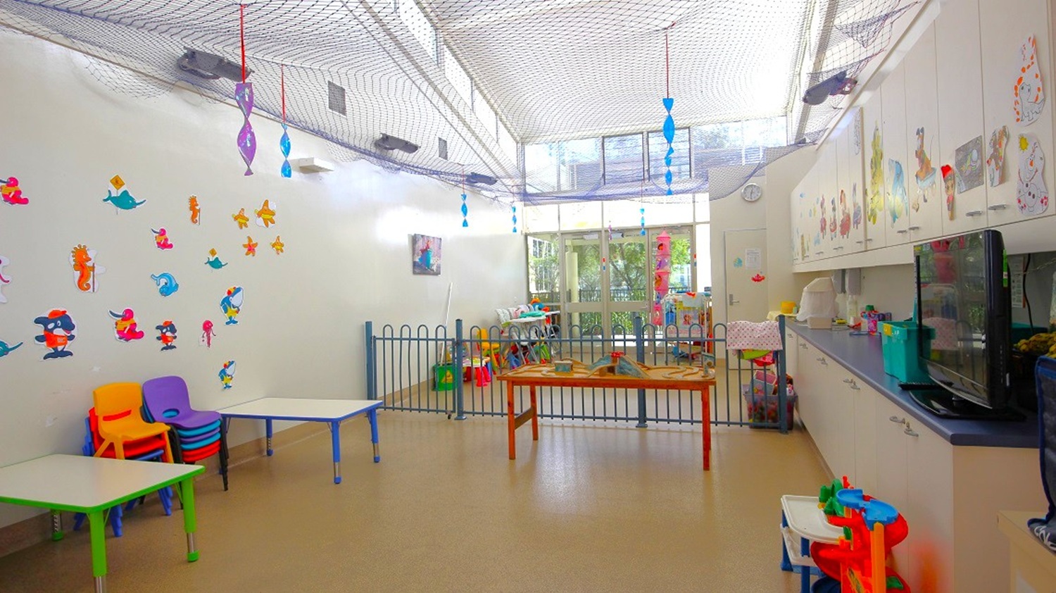 A brightly lit children's playroom with a high ceiling and netting. On the left, colourful chairs and tables are stacked, with cartoon character stickers on the wall. In the centre, a small fenced area contains toys and a table. The right side features a long counter with storage cabinets above and various items on top. The floor is light-coloured, and large windows in the background let in natural light. Hanging decorations are suspended from the ceiling.