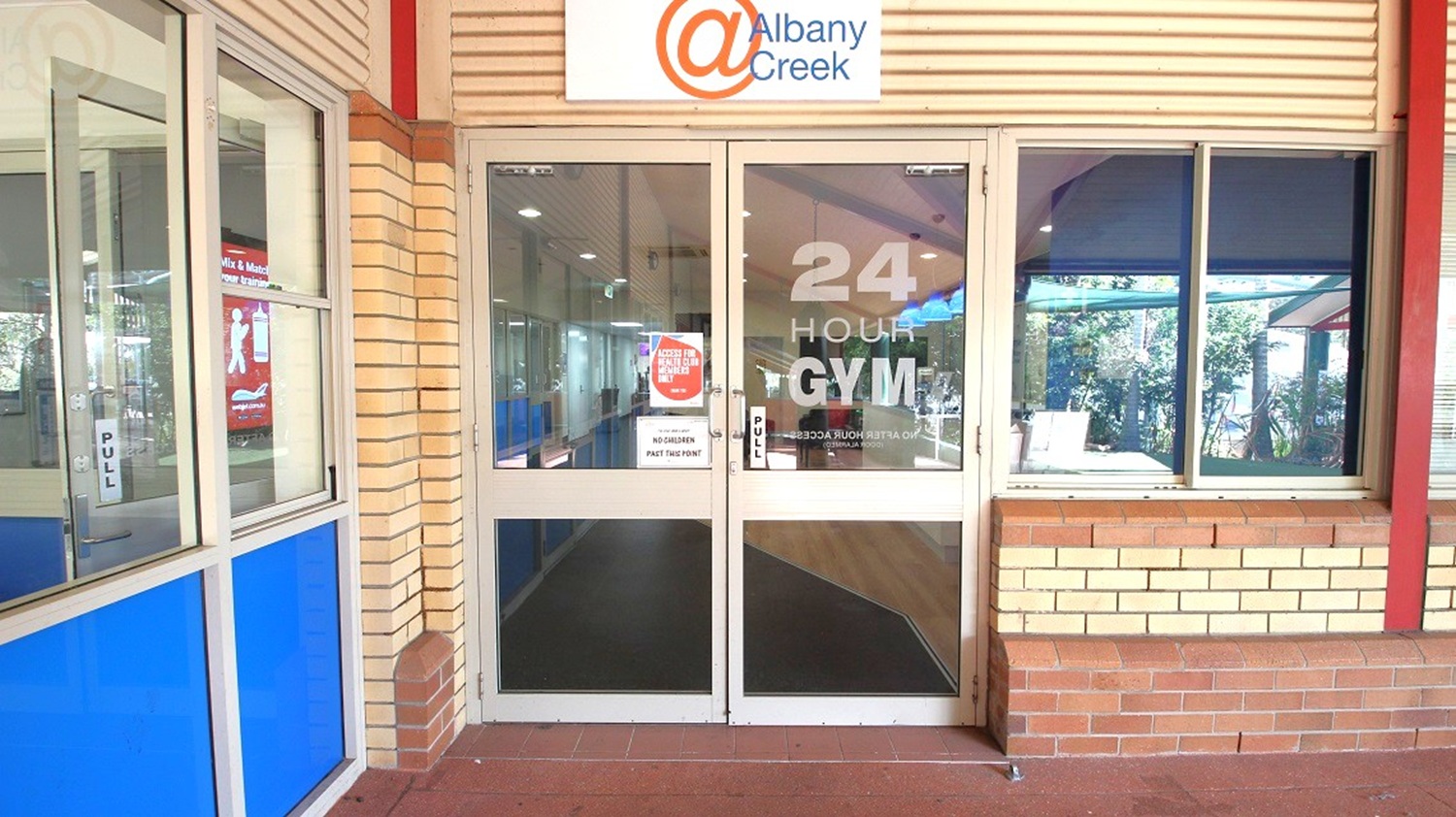 A glass entrance to a 24-hour gym with a sign above reading "@ Albany Creek". The doors have "24 HOUR GYM" written on them, and there are notices posted. To the left, a window with blue panels and a "PULL" sign. The building exterior is made of light-coloured bricks. On the right, a window shows a glimpse of greenery outside. The floor is tiled in a reddish-brown colour.