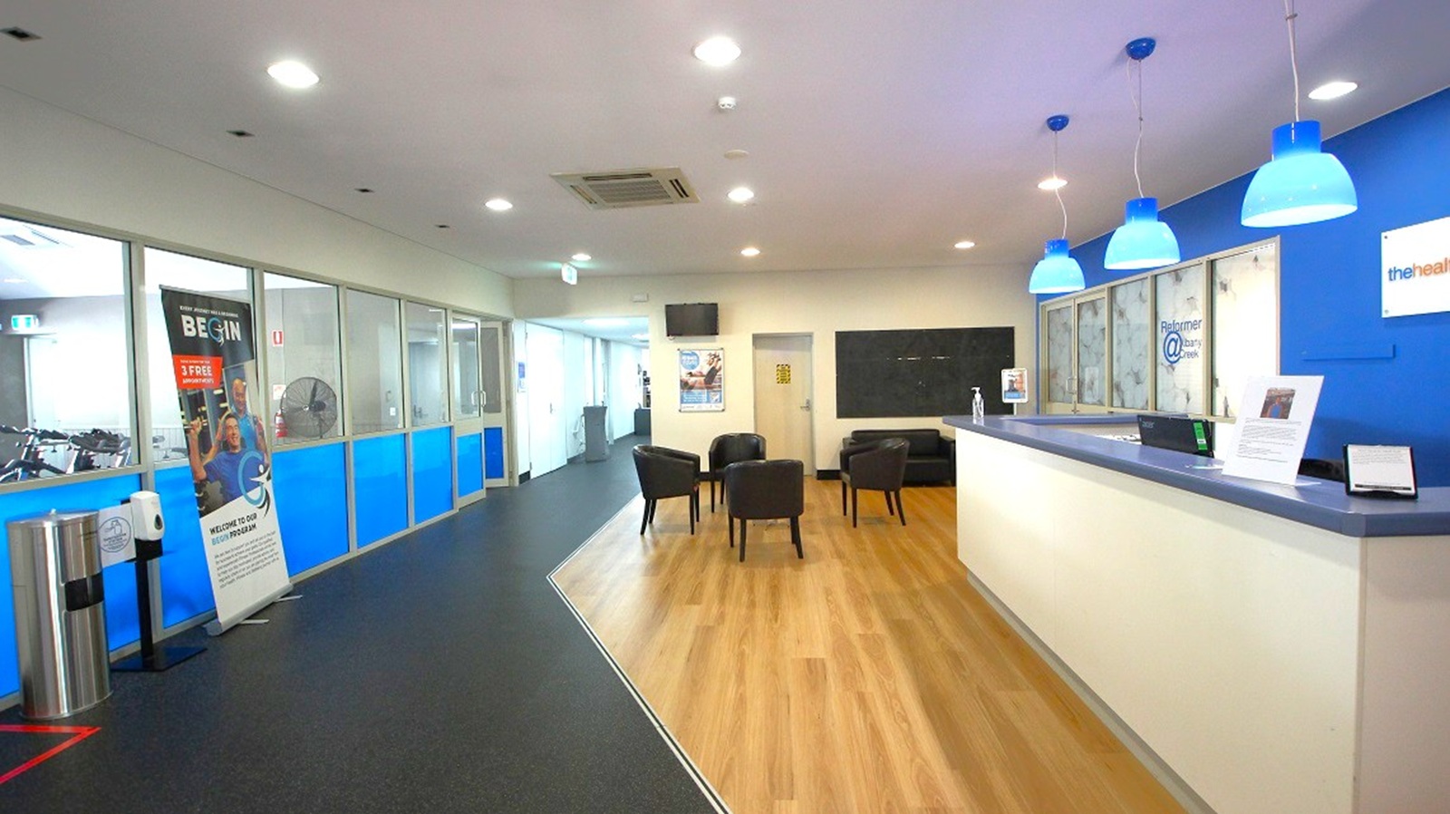 A modern reception area with a wooden floor. On the right, there's a long white reception desk with a dark countertop, illuminated by blue pendant lights. Behind the desk, a blue wall displays a sign. In the middle, several black chairs are arranged in a waiting area. On the left, a corridor with glass partitions and blue panels leads to other rooms. A poster and a hand sanitiser stand are near the entrance. The ceiling is white with recessed lighting.