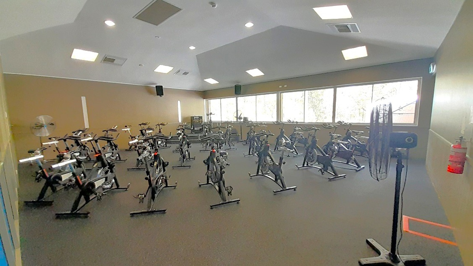 A room with multiple stationary bikes arranged in rows on a dark carpeted floor. Large windows on the right wall allow natural light to enter, with trees visible outside. A large fan is positioned on the right side near a fire extinguisher mounted on the wall. The ceiling is high with several recessed lights and air vents. The walls are painted in a neutral tone, and there are speakers mounted on the walls.