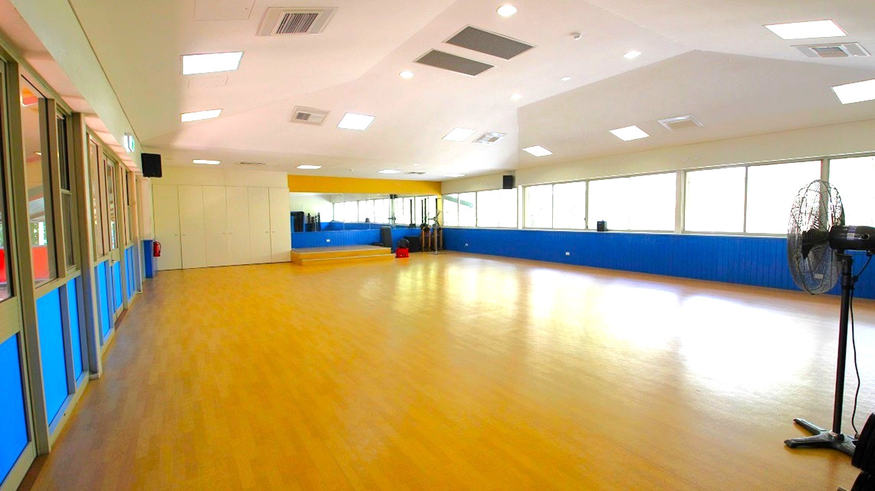 A spacious room with a wooden floor and a high ceiling featuring multiple recessed lights. On the left, there are large windows with green frames and blue lower panels. The right side has a row of windows allowing natural light to enter, with a fan visible in the foreground. At the far end, there's a small stage area with mirrors and exercise equipment. The walls are painted blue and white, and the ceiling has several ventilation units.