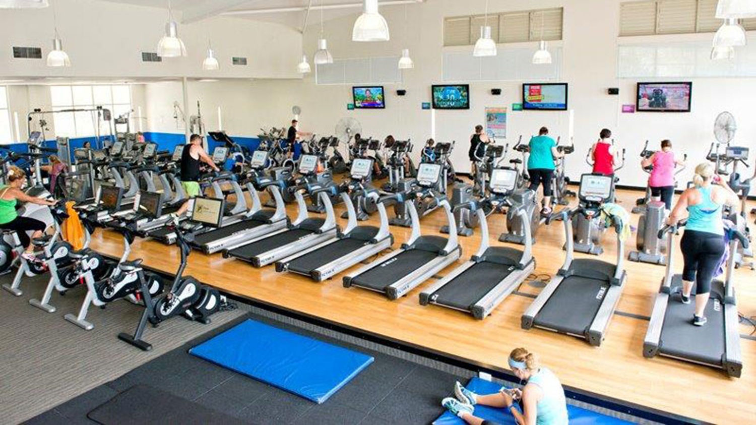 A spacious gym with a wooden floor and white ceiling. In the foreground, a person is stretching on a blue mat. The middle area features a row of treadmills and elliptical machines, with people exercising on them. The background has several wall-mounted televisions displaying various programmes. The gym is well-lit with hanging lights, and the walls are painted white with a blue accent.