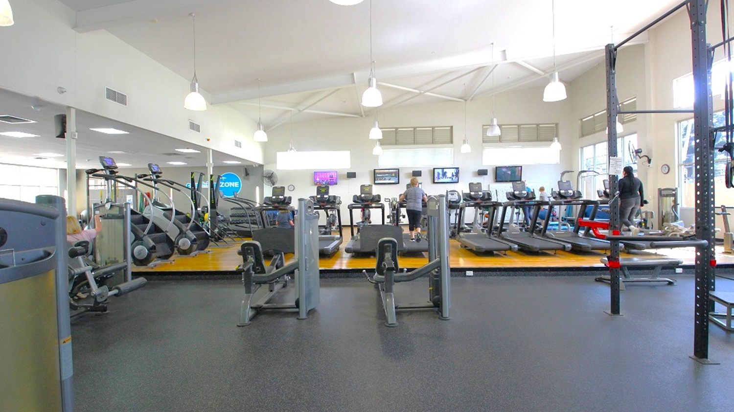 A spacious gym interior with a variety of exercise equipment. In the centre, there are several treadmills and elliptical machines on a raised wooden platform. To the left, more cardio machines are visible. On the right, a person is using a piece of equipment. The ceiling is high with white pendant lights, and the floor is covered with dark rubber mats. Large windows on the right allow natural light to enter, and several wall-mounted TVs are visible in the background.