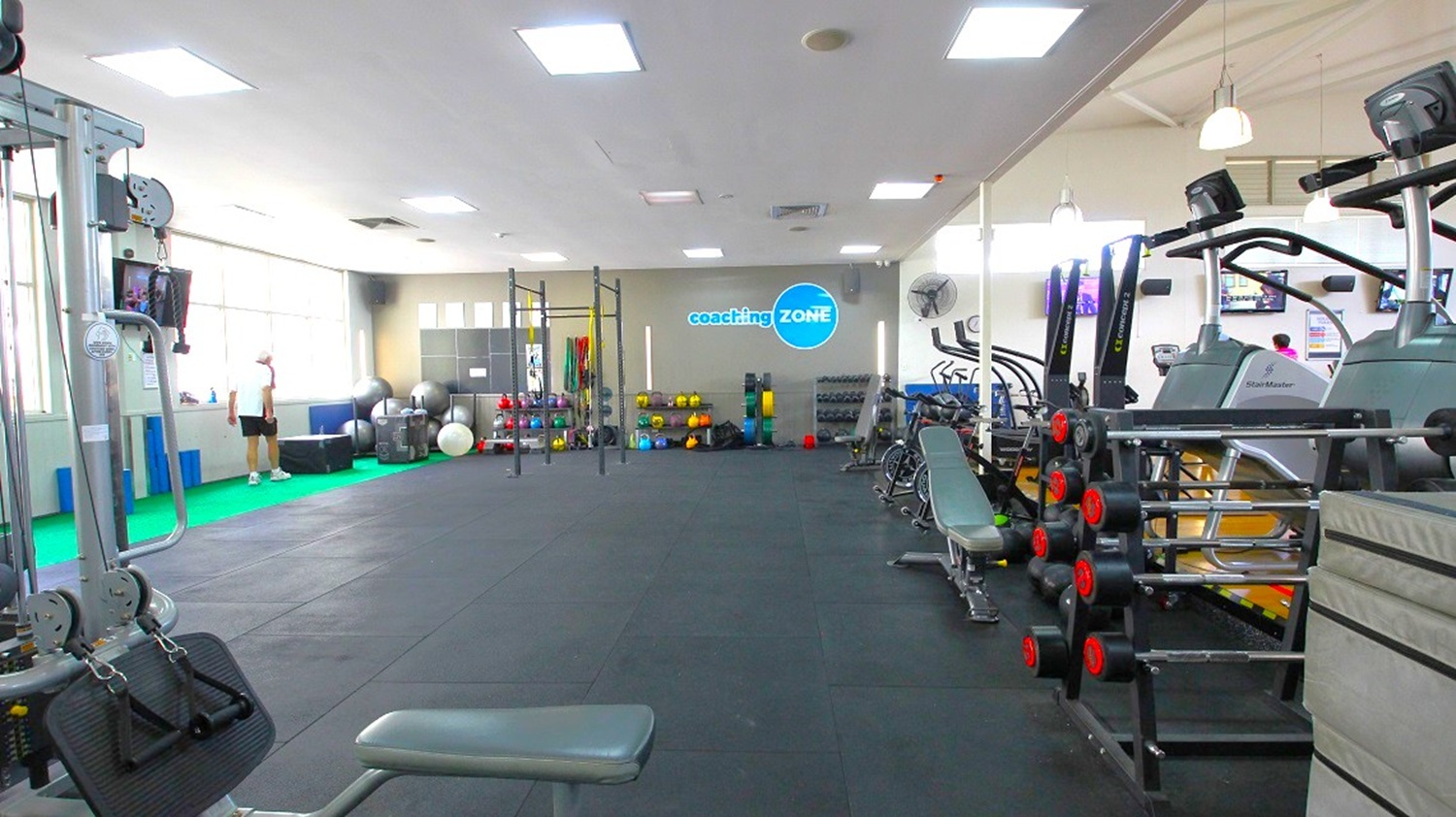 A spacious gym with a black rubber floor and white ceiling with recessed lighting. On the left, there are large windows and a person near exercise balls and equipment. In the centre, a rack with various weights and a sign reading "coaching ZONE" on the wall. On the right, treadmills and benches are lined up, with a rack of dumbbells nearby. The gym is well-lit and organised, with various fitness equipment throughout.