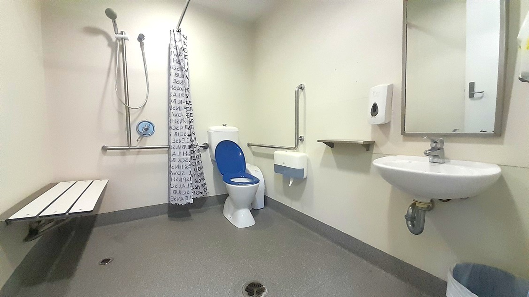 A bathroom with accessibility features. On the left, there's a shower area with a handheld showerhead and a white bench. A shower curtain with a pattern separates the shower from the rest of the room. In the centre, a toilet with a blue seat is equipped with grab bars. On the right, a wall-mounted sink with a mirror above it is visible, along with a soap dispenser and a small shelf. The floor is grey, and the walls are light-coloured.