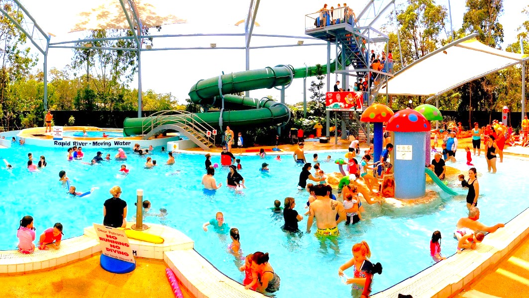 A busy indoor water park with a large pool filled with people of all ages. On the right, a tall green water slide with a staircase is visible, and a white canopy provides shade. Colourful mushroom-shaped water features are in the pool. On the left, a sign reads "Rapid River - Moving Water." The background has trees and a metal framework supporting the roof. The pool area is surrounded by a tiled deck.