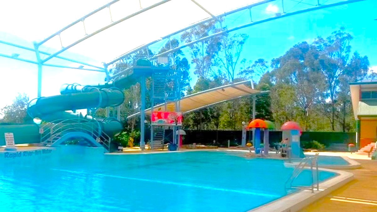 A swimming pool with a large, covered water slide on the left, featuring multiple twists and turns. The pool area is sheltered by a transparent roof structure. On the right, there are colourful water play features resembling mushrooms. In the background, tall trees are visible beyond the pool's perimeter. The pool deck is paved, and there is a building on the far right.