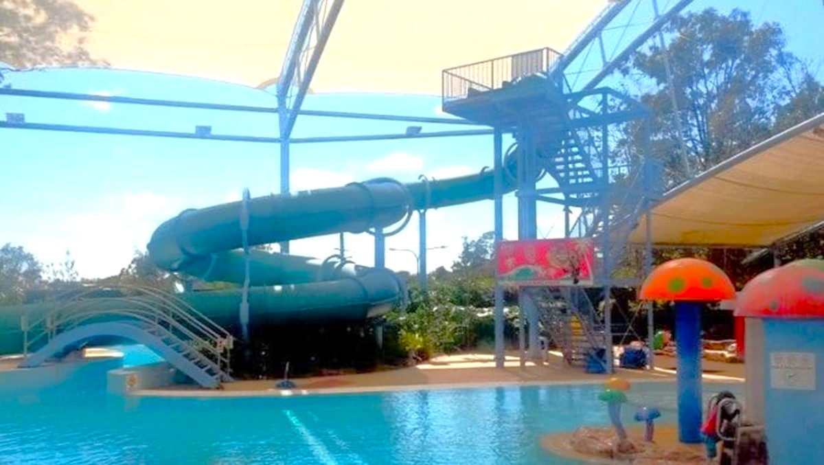 A water park with a large, twisting water slide on the left, supported by a metal structure. The slide ends in a pool of clear blue water. On the right, there are colourful mushroom-shaped water features. A canopy provides shade over part of the area. Trees and greenery are visible in the background, adding a natural element to the scene.