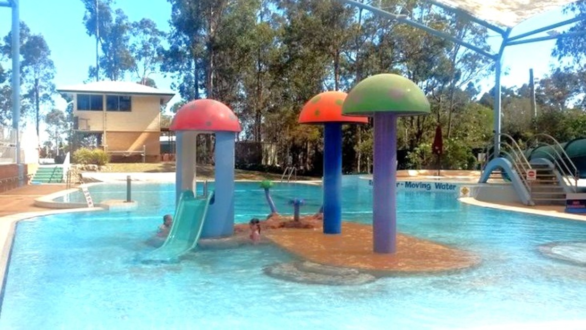 A children's pool area with colourful mushroom-shaped water features in the centre, each with a slide. The pool is shallow and surrounded by a paved area. In the background, there are trees and a building with a shaded roof. On the right, there is a larger slide with steps leading up to it. The area is covered by a large shade structure.