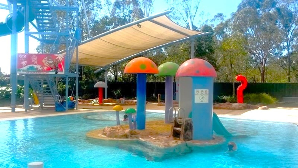 A children's water play area with a shallow pool. In the centre, colourful mushroom-shaped structures are positioned on a small island. To the left, a metal staircase leads to a water slide, with a large advertisement featuring ice cream. A shade canopy covers part of the area. In the background, there are trees and a red question mark-shaped structure. The pool's water is clear, and the surrounding area is paved.