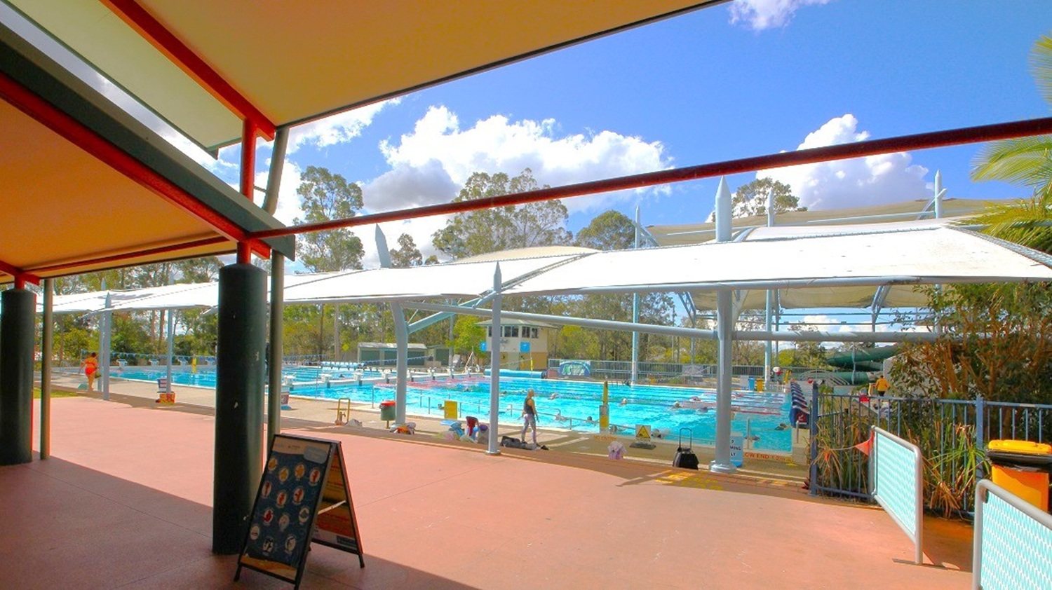 A covered outdoor swimming pool area with a large white canopy overhead. The pool is surrounded by trees and greenery in the background. In the foreground, there is a shaded walkway with a dark floor and ceiling, supported by black columns. A signboard is visible near the entrance, and a few people are around the pool area. The sky is partly cloudy.