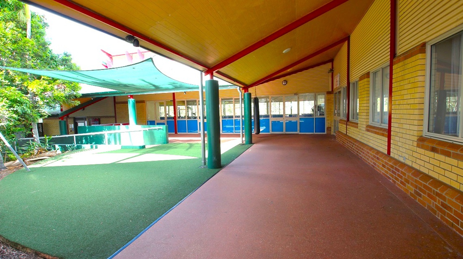 A covered outdoor area with a sloped roof supported by red beams and green columns. The floor is a mix of green and brown surfaces. On the left, there is a green shade sail above a play area, surrounded by trees. On the right, a brick wall with several windows. In the background, blue doors and windows line the building. The ceiling is a light brown colour.