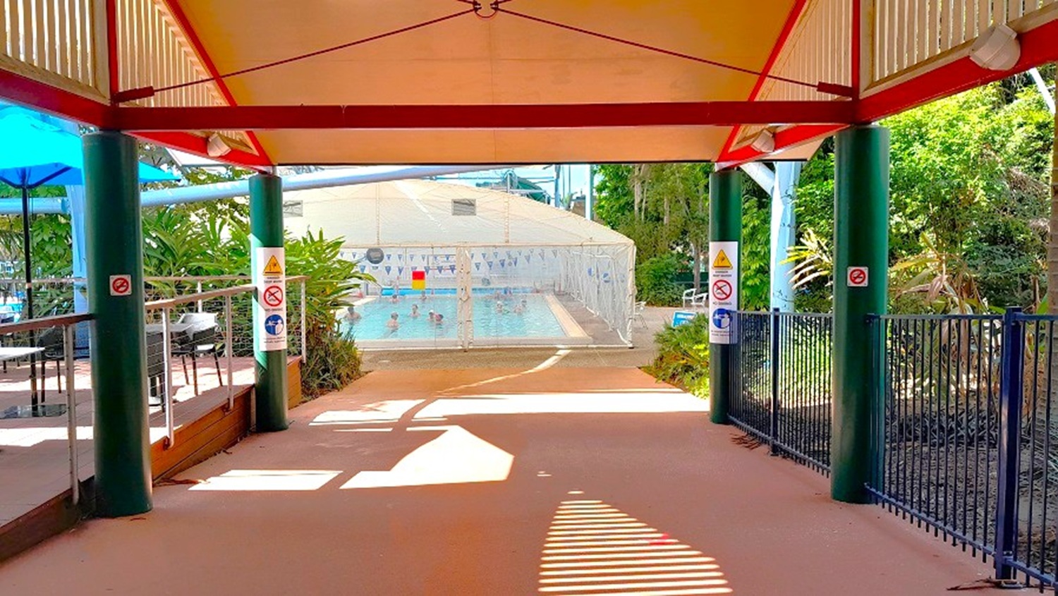 A covered walkway with a red and beige roof leads to a swimming pool area. The floor is a reddish-brown concrete. On the left, there are tables and chairs under blue umbrellas, bordered by a metal railing. On the right, a black metal fence and green foliage are visible. Signs with safety instructions are posted on the green pillars supporting the roof. In the background, the pool is enclosed by a white fence, with trees and plants surrounding the area.