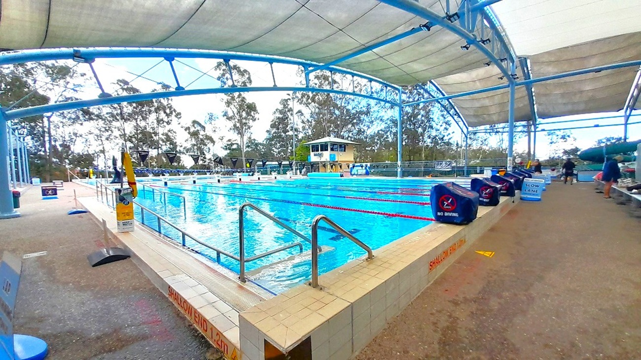 An outdoor swimming pool with multiple lanes, covered by a large canopy supported by blue metal beams. The pool is surrounded by a tiled deck with metal handrails leading into the water. On the right, there are timing devices and benches along the poolside. In the background, tall trees are visible beyond the pool area. The ground is a mix of tiles and concrete, and the ceiling is a white fabric stretched across the metal frame.