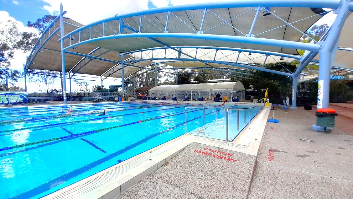 An outdoor swimming pool with multiple lanes, covered by a large, curved canopy supported by blue metal beams. The pool is divided by lane ropes, and a few swimmers are visible in the water. On the right, a concrete pool deck with a caution sign near the edge. In the background, trees and a transparent structure with people inside. The area is well-lit, and the pool water is a clear blue.