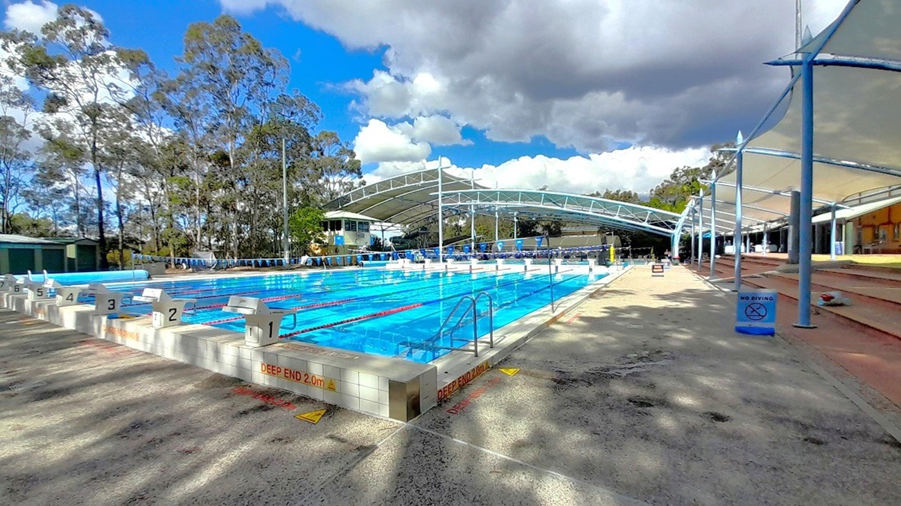 An outdoor swimming pool with multiple lanes, each marked with starting blocks numbered 1 to 8. The pool is surrounded by a paved area with a sign indicating a "Deep End." On the right, there is a covered seating area with tiered benches. A large canopy structure partially covers the pool, and trees are visible in the background. The sky is cloudy, casting shadows on the ground.