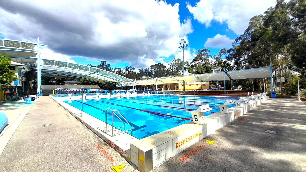 An outdoor swimming pool with multiple lanes, each marked with starting blocks numbered 1 to 8. The pool is surrounded by a tiled deck area with safety warnings. On the left, a covered seating area with a curved roof is visible. Trees and foliage line the background, and a partly cloudy sky is overhead.