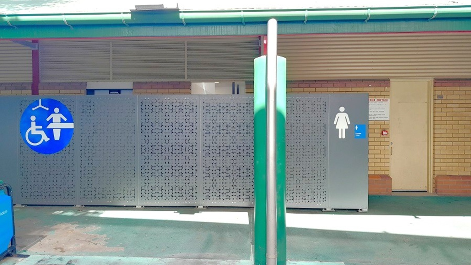 A public restroom area with a green pole in the foreground. On the left, there is a blue sign indicating accessible facilities with a baby changing symbol. The middle section has a decorative panel. On the right, a grey door with a female symbol and a small blue sign. The background features a beige wall with horizontal lines and a small doorway. The floor is concrete, and the ceiling has a green edge.