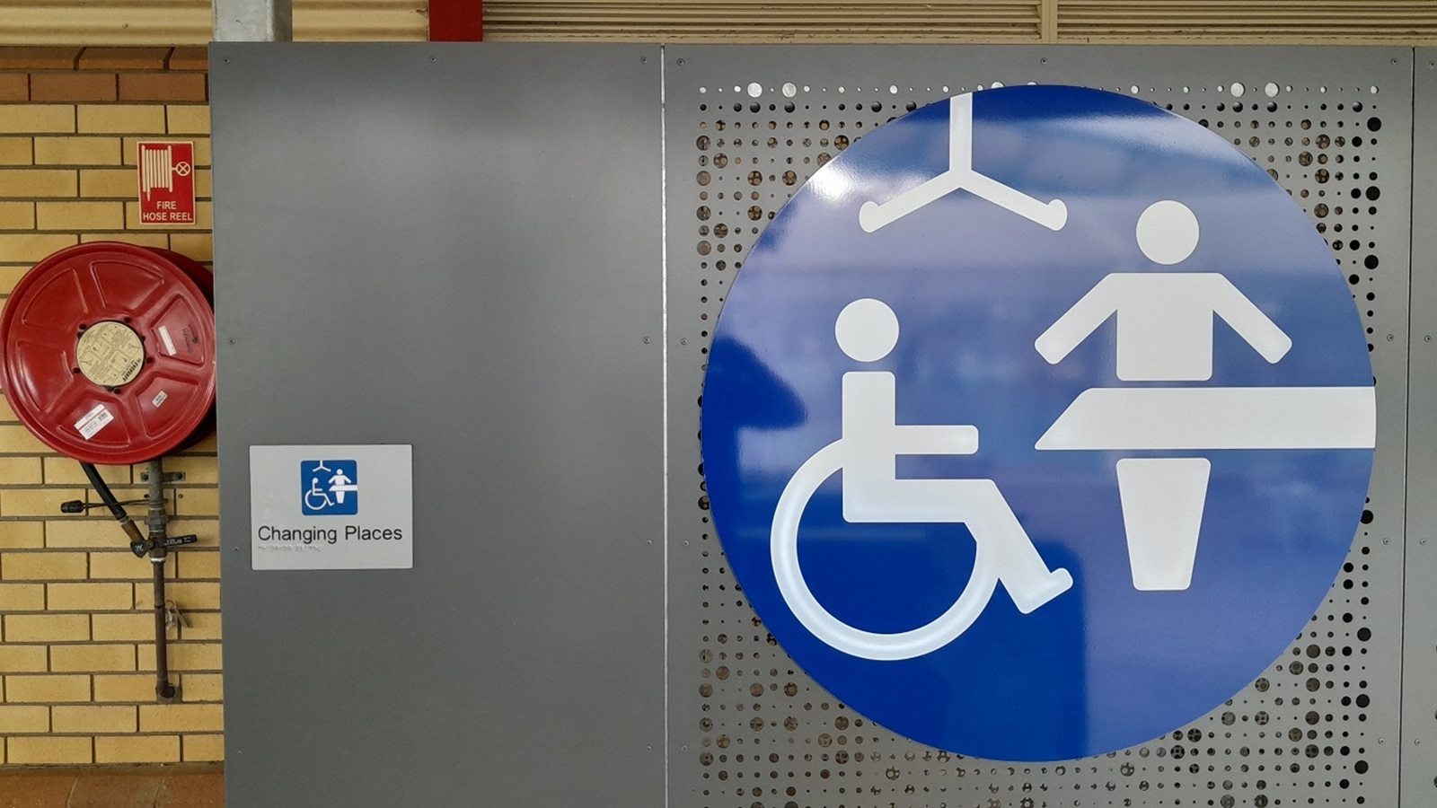 A large blue sign with white symbols indicating a wheelchair user and a person on a changing table is on the right. To the left, a red fire hose reel is mounted on a wall with light brown bricks. Below the sign, a smaller sign reads "Changing Places" with a similar symbol. The background features a perforated metal panel.