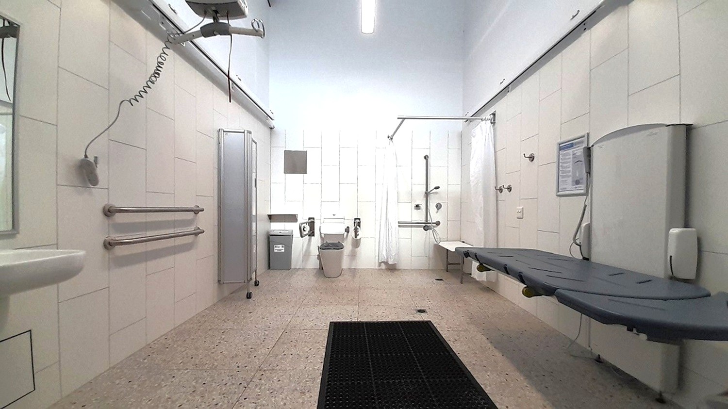 A spacious, accessible bathroom with a speckled floor and white walls. On the left, there are grab bars and a sink. In the centre, a toilet with support rails and a bin nearby. On the right, a shower area with a foldable seat and a long, adjustable bench. Overhead, a ceiling track hoist system is visible. The ceiling is plain with a fluorescent light fixture.