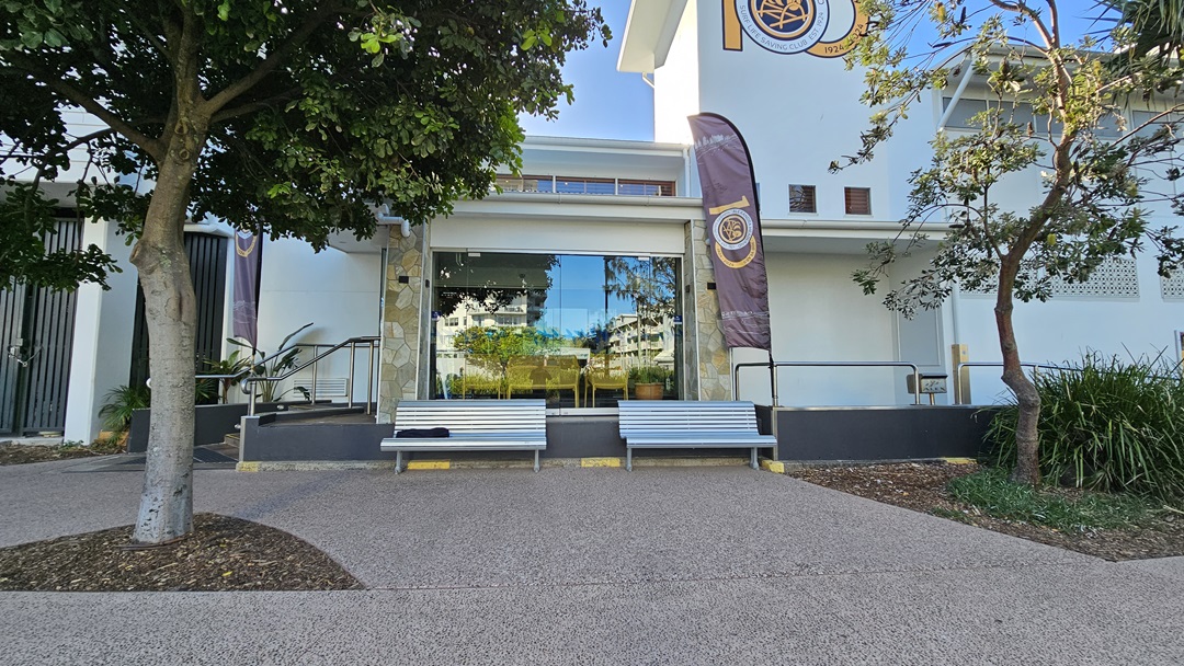 On the left, there is a tree with a thick trunk and green foliage. To the right, another tree with sparse leaves stands near a building. In the middle, a large glass window reflects the surrounding area, flanked by two grey benches with yellow accents. The background features a white building with a purple and gold flag. The floor is a textured, light brown pavement. On the left, there is a tree with a thick trunk and green foliage. To the right, another tree with sparse leaves stands near a building. In the middle, a large glass window reflects the surrounding area, flanked by two grey benches with yellow accents. The background features a white building with a purple and gold flag. The floor is a textured, light brown pavement.