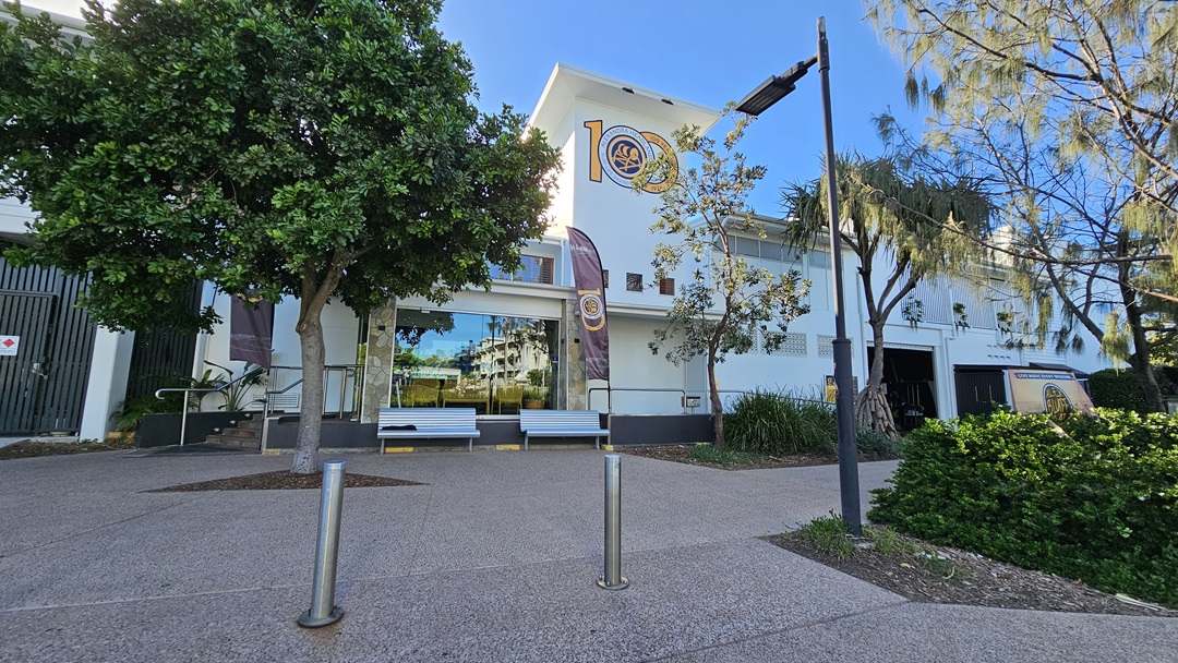 On the left, there is a large green tree next to a building with a glass entrance and a ramp. In the middle, two benches are placed in front of the entrance, flanked by two vertical banners. On the right, there is another tree with sparse foliage and a bushy area. The background features a white building with multiple windows and a sign. The floor is paved with a light brown, textured surface, and there is no visible ceiling. On the left, there is a large green tree next to a building with a glass entrance and a ramp. In the middle, two benches are placed in front of the entrance, flanked by two vertical banners. On the right, there is another tree with sparse foliage and a bushy area. The background features a white building with multiple windows and a sign. The floor is paved with a light brown, textured surface, and there is no visible ceiling.