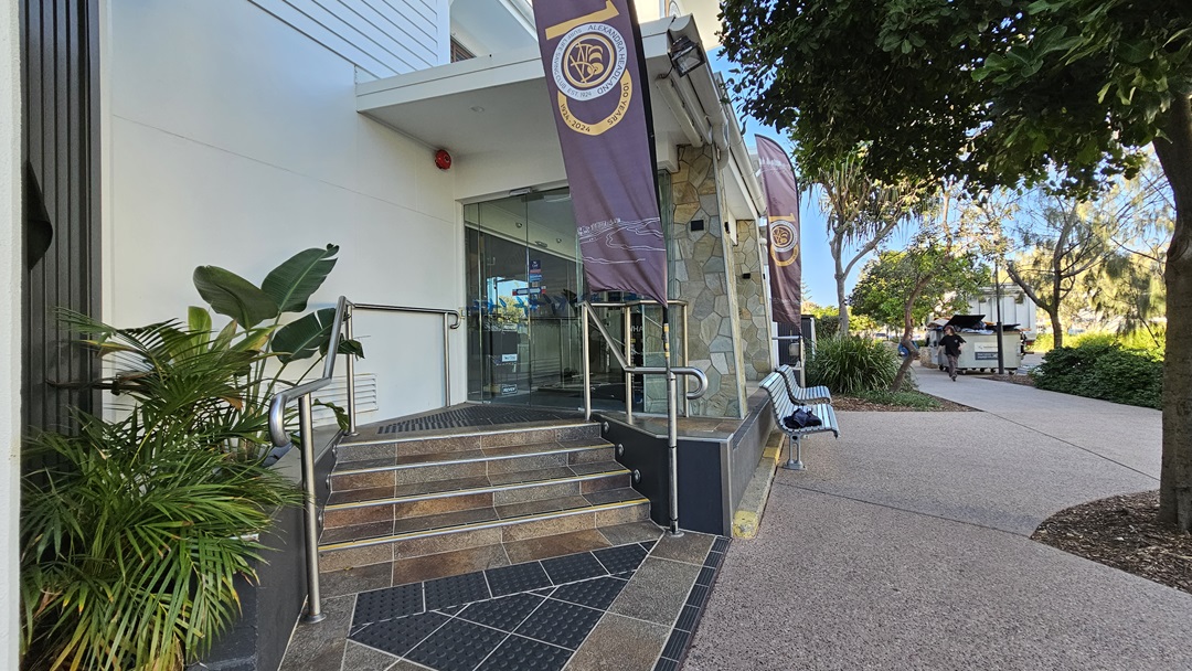 On the left, there are potted plants next to a building entrance with glass doors and a metal handrail. The middle features stone steps leading up to the entrance, flanked by two vertical banners. On the right, there is a bench and a tree with dense foliage. The background includes a pathway and more greenery. The floor is a mix of stone tiles and textured paving. On the left, there are potted plants next to a building entrance with glass doors and a metal handrail. The middle features stone steps leading up to the entrance, flanked by two vertical banners. On the right, there is a bench and a tree with dense foliage. The background includes a pathway and more greenery. The floor is a mix of stone tiles and textured paving.