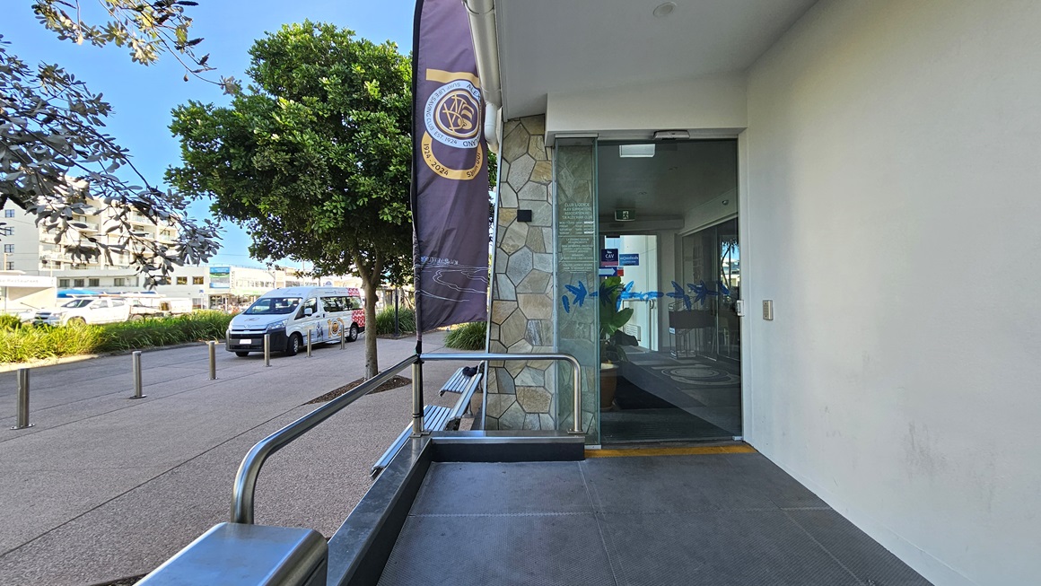 On the left, there is a tree with dense foliage next to a pathway where several vehicles are parked. In the middle, a vertical banner is attached to a pole near a stone wall. On the right, there is a glass door entrance with a metal handrail and a small bench beside it. The background includes more trees and buildings. The floor is a mix of concrete paving and a textured path, and the ceiling above the entrance is white with recessed lighting. On the left, there is a tree with dense foliage next to a pathway where several vehicles are parked. In the middle, a vertical banner is attached to a pole near a stone wall. On the right, there is a glass door entrance with a metal handrail and a small bench beside it. The background includes more trees and buildings. The floor is a mix of concrete paving and a textured path, and the ceiling above the entrance is white with recessed lighting.