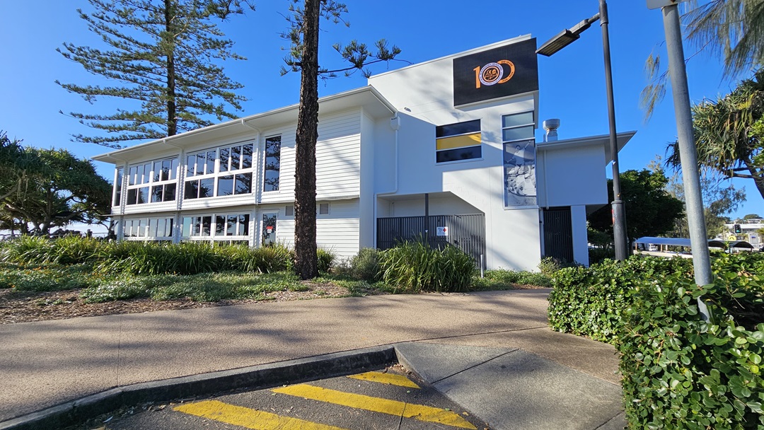 On the left, there is a large white building with multiple windows and a tree in front of it. In the middle, a tall tree stands next to a pathway bordered by grass and shrubs. On the right, there is a section of the building with a sign displaying the number "100" and a lamp post. The background includes more trees and greenery. The floor is a mix of concrete paving and a textured path with yellow markings. On the left, there is a large white building with multiple windows and a tree in front of it. In the middle, a tall tree stands next to a pathway bordered by grass and shrubs. On the right, there is a section of the building with a sign displaying the number "100" and a lamp post. The background includes more trees and greenery. The floor is a mix of concrete paving and a textured path with yellow markings.