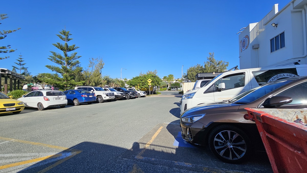 On the left, there are several parked cars, including a yellow one, next to a row of trees and bushes. In the middle, a road stretches into the distance with a pedestrian crossing sign visible. On the right, more parked cars are adjacent to a white building with a sign and a large window. The foreground features a textured asphalt surface with yellow markings. On the left, there are several parked cars, including a yellow one, next to a row of trees and bushes. In the middle, a road stretches into the distance with a pedestrian crossing sign visible. On the right, more parked cars are adjacent to a white building with a sign and a large window. The foreground features a textured asphalt surface with yellow markings.