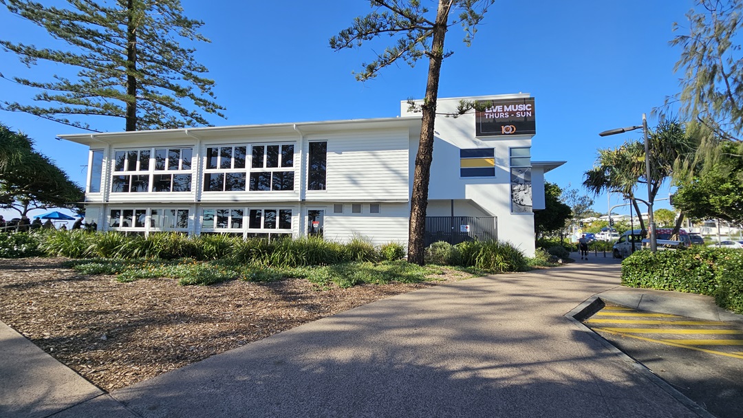 On the left, there is a white building with large windows and a garden area with mulch and low plants in front. In the middle, a tall tree stands next to the building. On the right, there is a pathway bordered by bushes and trees, leading to another part of the building with a sign that reads "LIVE MUSIC THURS - SUN" and the number "10". The floor is a mix of concrete paving and a textured path with yellow markings. On the left, there is a white building with large windows and a garden area with mulch and low plants in front. In the middle, a tall tree stands next to the building. On the right, there is a pathway bordered by bushes and trees, leading to another part of the building with a sign that reads "LIVE MUSIC THURS - SUN" and the number "10". The floor is a mix of concrete paving and a textured path with yellow markings.