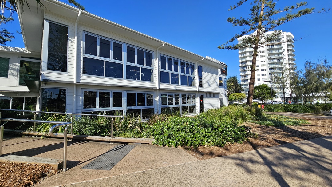 On the left, there is a white building with large windows and a metal handrail leading to a ramp. In the middle, there are bushes and plants in front of the building. On the right, a tall white apartment building is visible in the background, with trees and a pathway in the foreground. The floor is a mix of concrete paving and textured paths. On the left, there is a white building with large windows and a metal handrail leading to a ramp. In the middle, there are bushes and plants in front of the building. On the right, a tall white apartment building is visible in the background, with trees and a pathway in the foreground. The floor is a mix of concrete paving and textured paths.