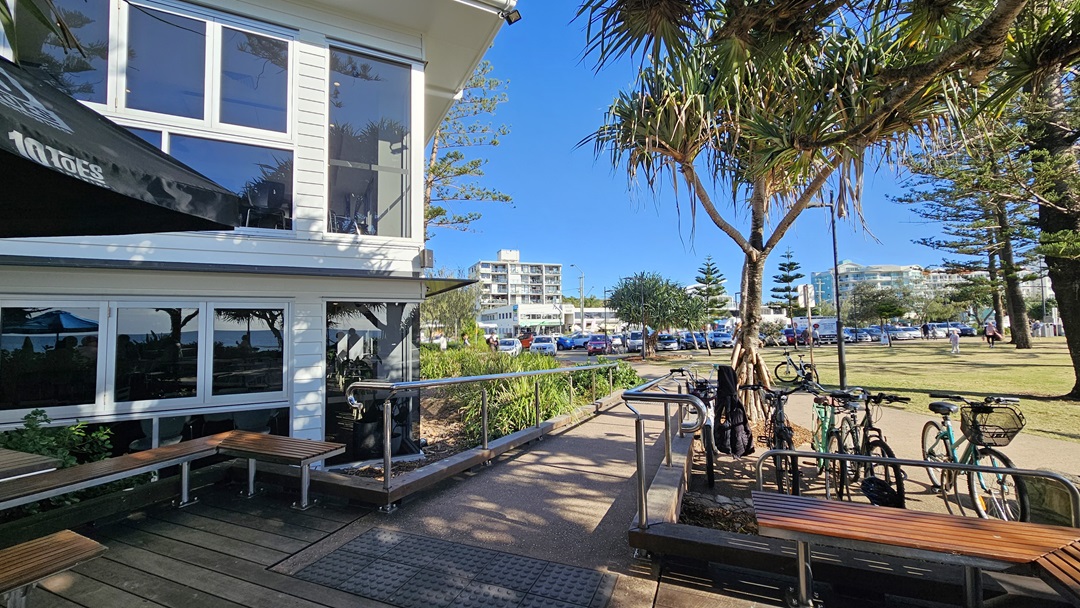 On the left, there is a white building with large windows and a black awning, next to a wooden bench and a metal handrail leading to a ramp. In the middle, there are several trees and a grassy area with parked bicycles. On the right, more trees and a pathway lead to a distant white apartment building. The foreground features a mix of wooden decking and concrete paving. On the left, there is a white building with large windows and a black awning, next to a wooden bench and a metal handrail leading to a ramp. In the middle, there are several trees and a grassy area with parked bicycles. On the right, more trees and a pathway lead to a distant white apartment building. The foreground features a mix of wooden decking and concrete paving.