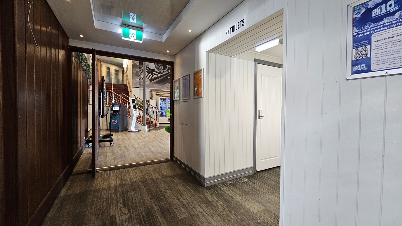On the left, there is a wooden panelled wall leading to an open area with a staircase and a surfboard display. In the middle, there is a hallway with a wooden floor and a green exit sign on the ceiling. On the right, there is a white wall with a door labelled "Toilets" and a blue poster beside it. The floor is a mix of wooden and carpeted areas, and the ceiling has recessed lighting and an exit sign. On the left, there is a wooden panelled wall leading to an open area with a staircase and a surfboard display. In the middle, there is a hallway with a wooden floor and a green exit sign on the ceiling. On the right, there is a white wall with a door labelled "Toilets" and a blue poster beside it. The floor is a mix of wooden and carpeted areas, and the ceiling has recessed lighting and an exit sign.
