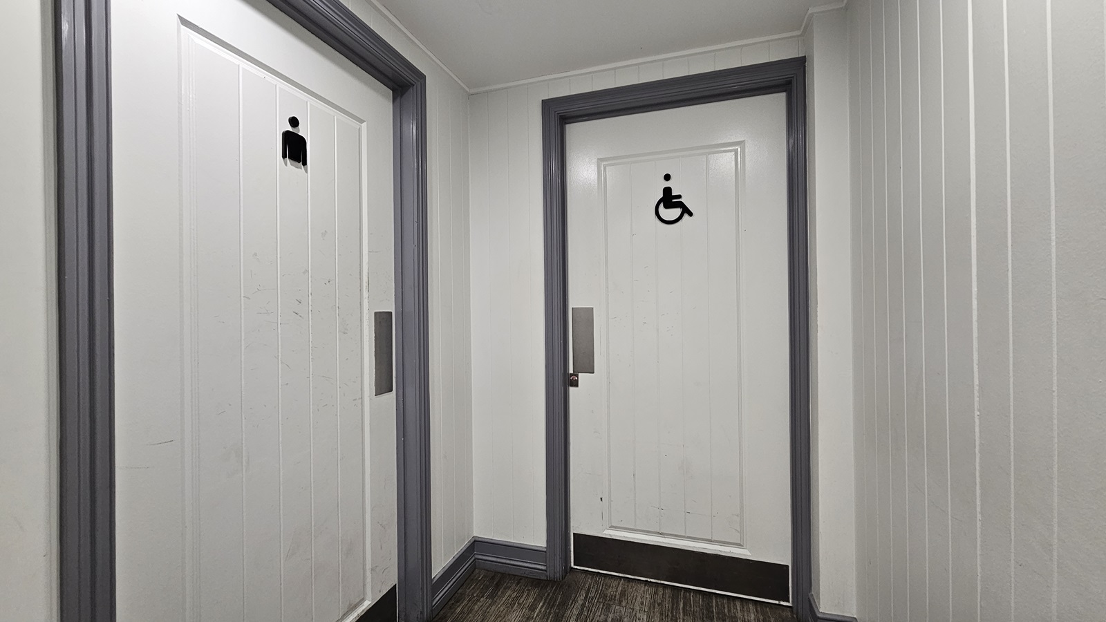 On the left, there is a white door with a male restroom symbol. In the middle, there is a white door with a wheelchair accessible symbol. Both doors have grey frames. On the right, there is a white panelled wall. The floor is dark wood, and the ceiling is white with recessed lighting. On the left, there is a white door with a male restroom symbol. In the middle, there is a white door with a wheelchair accessible symbol. Both doors have grey frames. On the right, there is a white panelled wall. The floor is dark wood, and the ceiling is white with recessed lighting.
