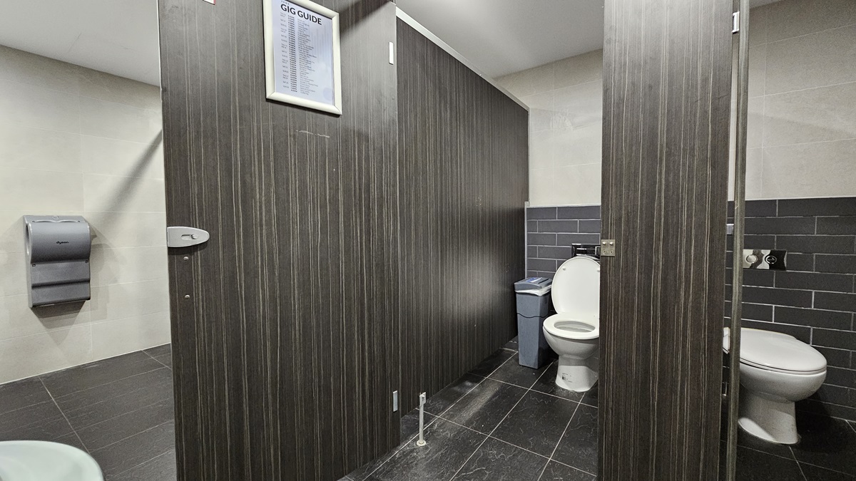 On the left, there is a grey paper towel dispenser mounted on a beige tiled wall. In the middle, there are two dark wood bathroom stalls, one with a closed door and the other open, revealing a white toilet and a blue bin. On the right, there is another white toilet with a grey tiled wall behind it. The floor is covered in dark grey tiles, and the ceiling is white with recessed lighting. On the left, there is a grey paper towel dispenser mounted on a beige tiled wall. In the middle, there are two dark wood bathroom stalls, one with a closed door and the other open, revealing a white toilet and a blue bin. On the right, there is another white toilet with a grey tiled wall behind it. The floor is covered in dark grey tiles, and the ceiling is white with recessed lighting.