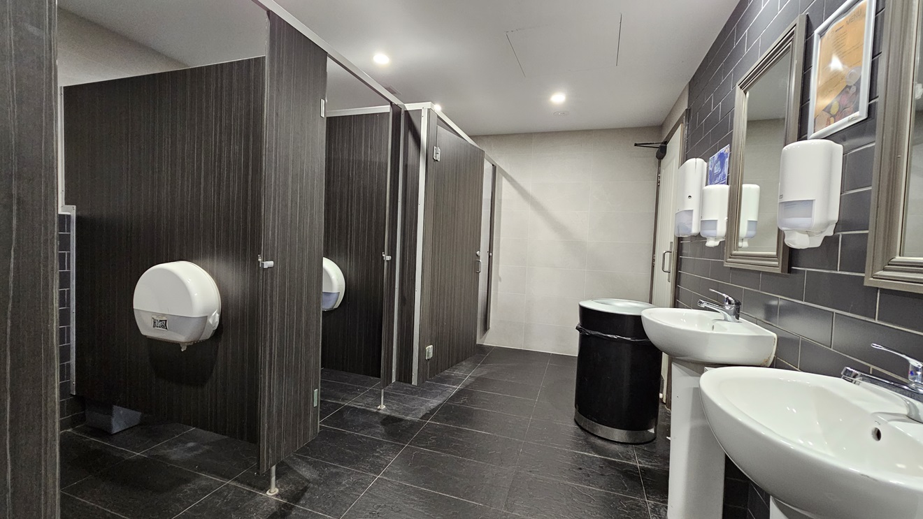 On the left, there are three dark wood bathroom stalls with closed doors, each with a white toilet paper dispenser. In the middle, there is a black bin against a beige tiled wall. On the right, there are two white sinks with mirrors above them, and three white soap dispensers mounted on a grey tiled wall. The floor is covered in dark grey tiles, and the ceiling is white with recessed lighting. On the left, there are three dark wood bathroom stalls with closed doors, each with a white toilet paper dispenser. In the middle, there is a black bin against a beige tiled wall. On the right, there are two white sinks with mirrors above them, and three white soap dispensers mounted on a grey tiled wall. The floor is covered in dark grey tiles, and the ceiling is white with recessed lighting.
