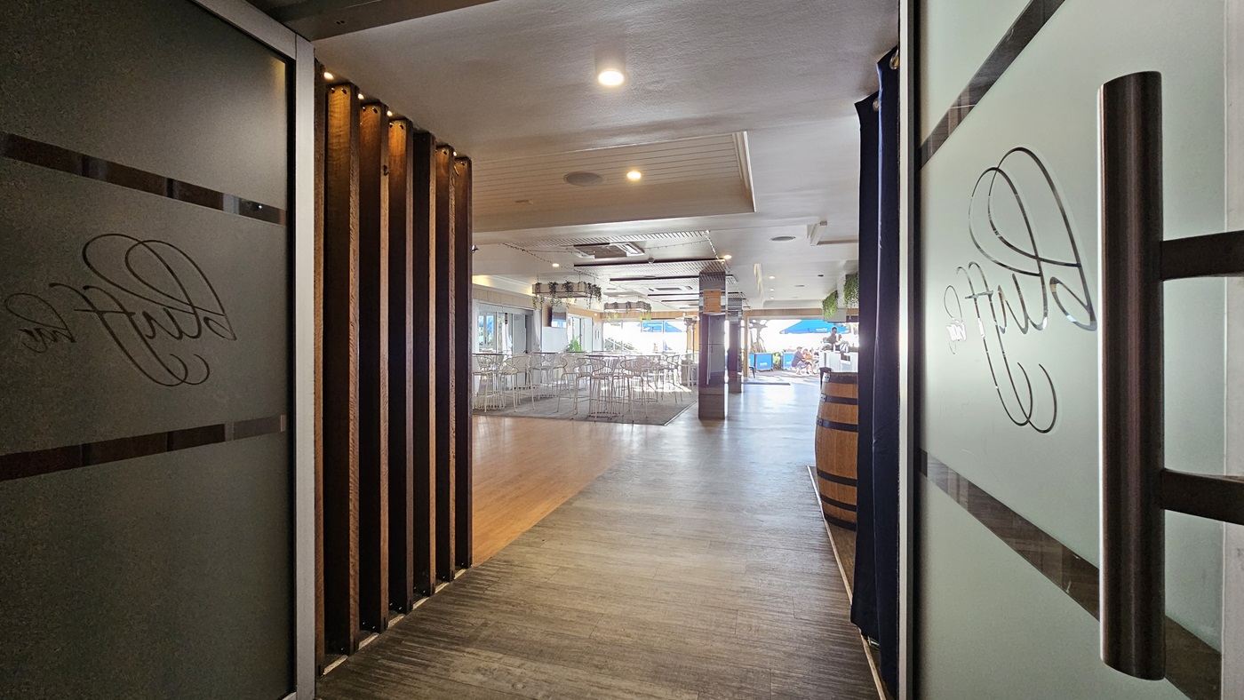 On the left, there is a frosted glass door with the word "Bluff" etched on it, partially open, revealing a wooden panelled wall. In the middle, there is a hallway with a wooden floor leading to a dining area with tables and chairs. In the background, there is a bar area with a wooden barrel and a blue umbrella visible through the windows. On the right, there is another frosted glass door with the word "Bluff" etched on it, also partially open. The ceiling is white with recessed lighting. On the left, there is a frosted glass door with the word "Bluff" etched on it, partially open, revealing a wooden panelled wall. In the middle, there is a hallway with a wooden floor leading to a dining area with tables and chairs. In the background, there is a bar area with a wooden barrel and a blue umbrella visible through the windows. On the right, there is another frosted glass door with the word "Bluff" etched on it, also partially open. The ceiling is white with recessed lighting.