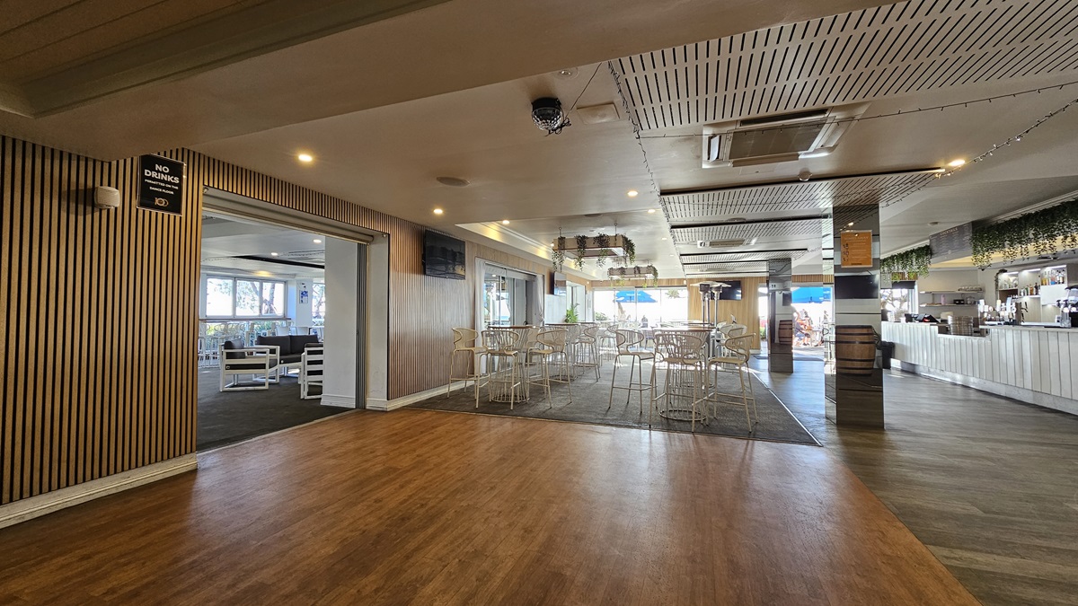 On the left, there is a wooden panelled wall with a "No Smoking" sign and an open doorway leading to a seating area with white chairs and tables. In the middle, there is a dining area with white tables and chairs on a dark carpet. In the background, there are large windows with views of the outside and a bar area with a blue umbrella. On the right, there is a long white bar counter with bottles and glasses. The floor transitions from wood to carpet to tile, and the ceiling has recessed lighting. On the left, there is a wooden panelled wall with a "No Smoking" sign and an open doorway leading to a seating area with white chairs and tables. In the middle, there is a dining area with white tables and chairs on a dark carpet. In the background, there are large windows with views of the outside and a bar area with a blue umbrella. On the right, there is a long white bar counter with bottles and glasses. The floor transitions from wood to carpet to tile, and the ceiling has recessed lighting.