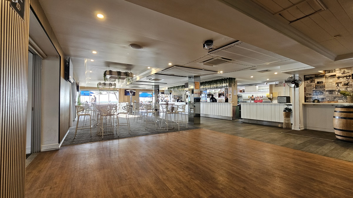 On the left, there is a wall with vertical blinds and a partially open sliding door. In the middle, there is a dining area with white metal chairs and tables on a dark carpet. In the background, there are large windows with views of the outside and blue umbrellas. On the right, there is a long white bar counter with bottles and glasses, a wooden barrel, and a wall with framed photos. The floor transitions from wood to carpet to tile, and the ceiling has recessed lighting. On the left, there is a wall with vertical blinds and a partially open sliding door. In the middle, there is a dining area with white metal chairs and tables on a dark carpet. In the background, there are large windows with views of the outside and blue umbrellas. On the right, there is a long white bar counter with bottles and glasses, a wooden barrel, and a wall with framed photos. The floor transitions from wood to carpet to tile, and the ceiling has recessed lighting.