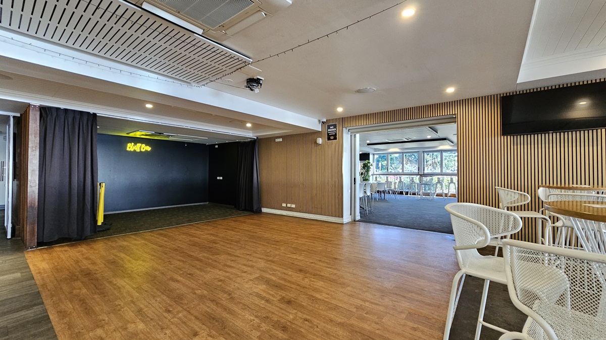 On the left, there is a black curtain partially covering a doorway with a neon "Bluff" sign above it. In the middle, there is an open space with a wooden floor leading to another room with large windows and white chairs. In the background, the room has a dark carpet and more white chairs. On the right, there is a wall-mounted TV above a wooden panelled wall and a seating area with white metal chairs and tables. The ceiling is white with recessed lighting and air conditioning units. On the left, there is a black curtain partially covering a doorway with a neon "Bluff" sign above it. In the middle, there is an open space with a wooden floor leading to another room with large windows and white chairs. In the background, the room has a dark carpet and more white chairs. On the right, there is a wall-mounted TV above a wooden panelled wall and a seating area with white metal chairs and tables. The ceiling is white with recessed lighting and air conditioning units.