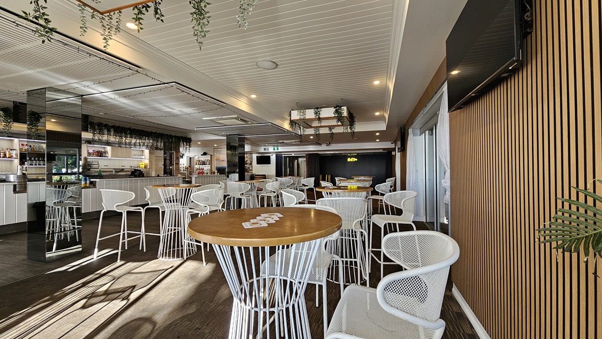 On the left, there are white cabinets and a bar area with shelves of bottles and glasses, decorated with hanging plants. In the middle, there are round wooden tables with white metal chairs on a dark carpet. In the background, there is a stage area with a black curtain and a neon "Bluff" sign. On the right, there is a wooden panelled wall with a wall-mounted TV and a sliding glass door. The floor is dark carpet, and the ceiling is white with recessed lighting and hanging plants. On the left, there are white cabinets and a bar area with shelves of bottles and glasses, decorated with hanging plants. In the middle, there are round wooden tables with white metal chairs on a dark carpet. In the background, there is a stage area with a black curtain and a neon "Bluff" sign. On the right, there is a wooden panelled wall with a wall-mounted TV and a sliding glass door. The floor is dark carpet, and the ceiling is white with recessed lighting and hanging plants.