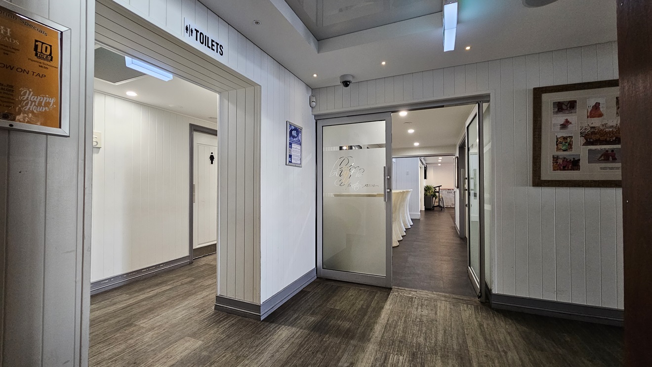 On the left, there is a doorway with a sign above it reading "Toilets," leading to a hallway with white doors. In the middle, there is a glass door with "Bluff" etched on it, opening into a room with white walls and a dark floor. In the background, there is a long table with a white tablecloth and a lit exit sign. On the right, there is a white panelled wall with a large framed collage of photos. The floor is dark wood, and the ceiling is white with recessed lighting. On the left, there is a doorway with a sign above it reading "Toilets," leading to a hallway with white doors. In the middle, there is a glass door with "Bluff" etched on it, opening into a room with white walls and a dark floor. In the background, there is a long table with a white tablecloth and a lit exit sign. On the right, there is a white panelled wall with a large framed collage of photos. The floor is dark wood, and the ceiling is white with recessed lighting.